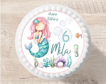 Puede incluir: Un pastel blanco con una imagen comestible con temática de sirena. La imagen presenta una sirena con cabello rosado y cola verde, un número 6, el nombre Mila y detalles de la vida marina. El texto dice "Feliz cumpleaños".