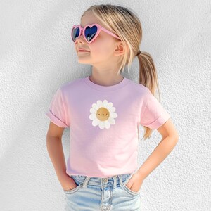 Puede incluir: Una camiseta rosa claro con un gr&aacute;fico de margarita sonriente. La camiseta la lleva una persona joven que lleva gafas de sol rosas en forma de coraz&oacute;n y vaqueros azules. Las mangas est&aacute;n dobladas. El fondo es una pared blanca.