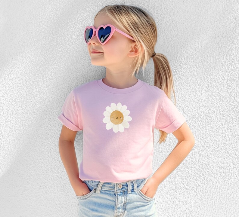 Puede incluir: Una camiseta rosa claro con un gr&aacute;fico de margarita sonriente. La camiseta la lleva una persona joven que lleva gafas de sol rosas en forma de coraz&oacute;n y vaqueros azules. Las mangas est&aacute;n dobladas. El fondo es una pared blanca.