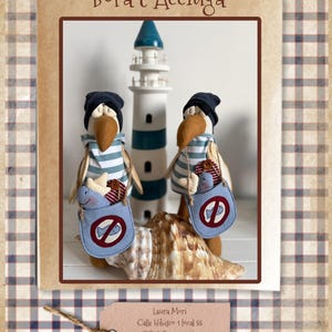May include: Two fabric bird figurines wearing dark blue knit caps and light blue and white striped shirts. Each bird carries a light blue pouch featuring a red circle with a diagonal line over a fish icon. A wooden lighthouse and a seashell are present.