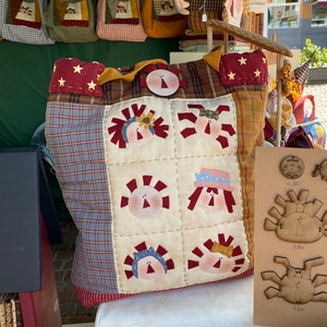 Può includere: Una borsa a tracolla in patchwork con un design a quadri rosso, bianco e blu. La borsa presenta una varietà di applicazioni, tra cui stelle, fiori e volti. La borsa è appoggiata su un tavolo con un cartello che dice "0,80" e "2,50".