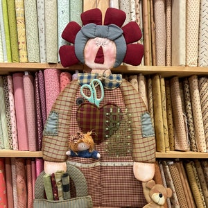 Può includere: Una bambola di patchwork rosso e bianco con una testa di fiore rosso e un orsacchiotto di peluche marrone. La bambola tiene un cestino con ritagli di stoffa e un righello. La bambola è in piedi davanti a uno scaffale di rotoli di stoffa.