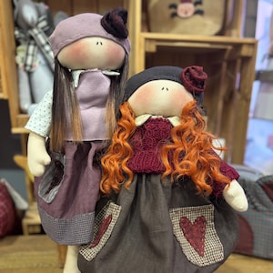 May include: Two handmade rag dolls with long hair. The doll on the left is wearing a purple dress with a white and purple checkered trim. The doll on the right is wearing a burgundy sweater and a brown skirt with a red heart patch and a red and white checkered trim.