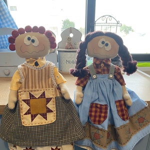 May include: Two handmade rag dolls with brown hair and big eyes. The doll on the left is wearing a yellow and brown plaid dress with a star-shaped apron. The doll on the right is wearing a blue and brown plaid dress with a blue and white gingham skirt.