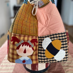 Puede incluir: Una bolsa de mano acolchada con un diseño de patchwork que presenta una niña, una abeja y patrones de cuadros. La bolsa tiene un cierre de cordón y asas de color turquesa con lunares dorados.
