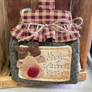Puede incluir: Una bolsa de tela de cuadros rojos y beige con una cara de reno de tela marrón y el texto "Magic Reindeer Kisses".