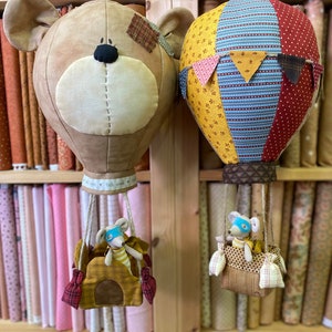 Puede incluir: Dos globos aerostáticos hechos a mano con diseños de patchwork de tela. Un globo tiene la forma de la cabeza de un oso y el otro tiene un patrón de rayas de colores. Cada globo tiene una pequeña figura de ratón en una cesta.