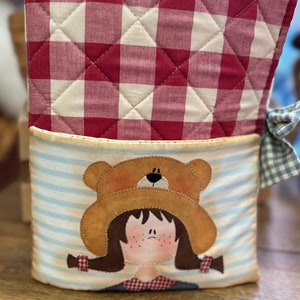 Puede incluir: Una bolsa de almacenamiento de tela acolchada con un patrón de cuadros rojos y blancos en la parte superior y un patrón de rayas azules y blancas en la parte inferior. La sección inferior presenta una aplicación de una niña con cabello castaño que lleva un sombrero marrón con una cara de oso en la parte superior.