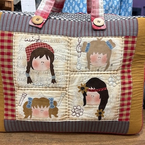 Op de afbeelding: Een gevoerde schoudertas met een rood-wit-blauw ruitpatroon. De tas heeft vier geborduurde afbeeldingen van meisjes met verschillende kapsels en accessoires. De tekst "I wish wearing fla on cald grating up brit lud will be oses and Kittines Caty" is geborduurd op de tas.