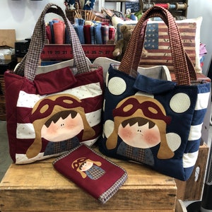 Peut inclure: Deux sacs cabas en tissu matelassé avec un motif de drapeau américain rouge, blanc et bleu. Chaque sac présente un personnage de dessin animé avec un chapeau d'aviateur brun et des lunettes. Les sacs sont posés sur une surface en bois avec une petite pochette en tissu rouge devant eux.