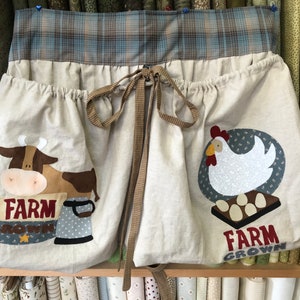 May include: A beige fabric storage bag with two pockets, each featuring a different farm animal design. The left pocket has a brown cow with the text "FARM GROWN" and the right pocket has a white chicken with the text "FARM GROWN".