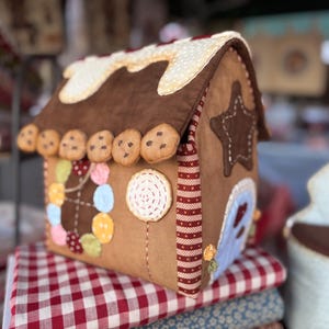 Puede incluir: Un juguete de peluche marrón con forma de casa de jengibre con un techo blanco, decorado con caramelos de colores, chispas de chocolate y un ribete rojo y blanco.