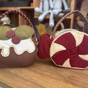 Può includere: Due cesti regalo di Natale in tessuto. Uno è a forma di bastoncino di zucchero con strisce rosse e bianche e l'altro è a forma di budino di Natale con tessuto marrone, verde e bianco.