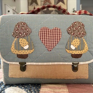 以下が含まれることがあります： 茶色と白のチェック柄の青いキルティングバッグ。バッグには茶色の布製のハンドルが2つと金属製の留め金が2つ付いています。バッグの前面には、茶色の帽子と白のドット柄のドレスを着た女の子のアップリケが2つ付いています。女の子たちの間に、赤と白のチェック柄のハートが中央に配置されています。