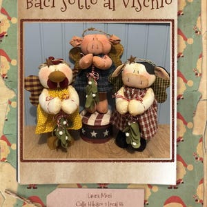 Puede incluir: Tres muñecas de ángel de tela hechas a mano: una gallina, un cerdo y una vaca. Las muñecas visten vestidos y sostienen muérdago. La muñeca de cerdo está sobre un pequeño soporte redondo con un diseño de estrellas y rayas. El texto "Baci sotto al vischio" está presente.