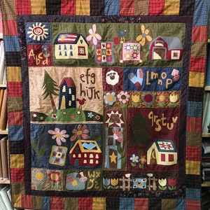 Può includere: Una trapunta colorata a patchwork con un tema campestre. La trapunta presenta case, alberi, fiori e l'alfabeto. La trapunta è realizzata con vari tessuti in tonalità di rosso, verde, blu, giallo e marrone.