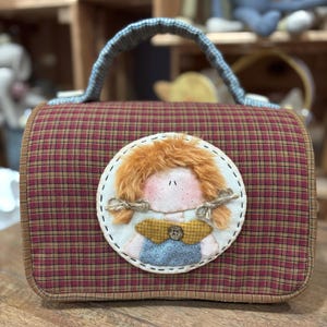 Può includere: Una borsa a mano in tessuto a quadri rosso e marrone con un'applicazione rotonda di una ragazza con i capelli rossi. La ragazza indossa un vestito blu con un colletto giallo e un bottone bianco.