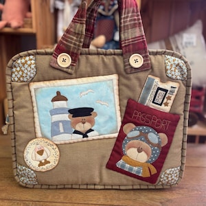 Peut inclure: Sac de voyage en tissu marron avec un motif de phare et d'ours. Le sac présente des appliqués de passeport et de billet, ainsi qu'un petit écusson de chien. Les poignées sont à carreaux et le sac est orné de boutons en bois.