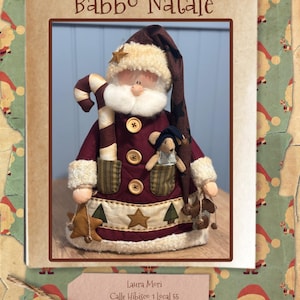 Puede incluir: Muñeco navideño artesanal con forma de Papá Noel, con un abrigo burdeos con ribetes color crema y un gorro a juego. El muñeco sostiene un bastón de caramelo y tiene un pequeño oso en el bolsillo. El texto "Babbo Natale" está en la parte superior.