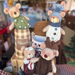 Puede incluir: Una colección de muñecos de oso de peluche hechos a mano, cada uno con atuendos y accesorios únicos. Un oso lleva un vestido verde con lunares blancos y una corona de estrella dorada. Otro está vestido de muñeco de nieve, y otros tienen atuendos festivos.