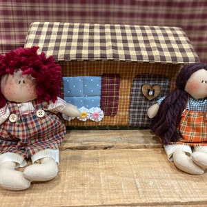 May include: Two handmade rag dolls sitting in front of a small house-shaped box. The dolls are dressed in plaid and have yarn hair. The house is decorated with plaid fabric and has a heart-shaped door.