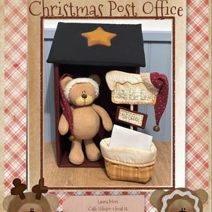 Puede incluir: Una exhibición navideña con un oso de peluche con un sombrero, junto a una oficina de correos en miniatura. La oficina de correos tiene un techo negro con una estrella dorada y un letrero que dice "North Pole Post Office". También se incluye una pequeña cesta.