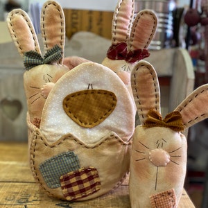 Może przedstawiać: Trzy ręcznie robione króliki wielkanocne z tkaniny, jeden w patchworkowym pojemniku w kształcie jajka. Króliki są beżowe z brązowymi oczami i haftowanymi detalami. Jajko jest beżowe z brązową, niebieską i czerwoną tkaniną patchworkową.