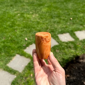 May include: A small, cylindrical object with a smooth, polished finish and a warm, amber-orange color featuring swirling, wood-grain-like patterns throughout its surface. The item has a flat top and a slightly tapered base, showcasing a glossy texture.