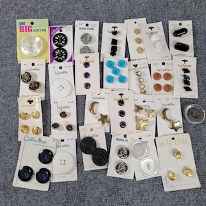 May include: A collection of various buttons, including black, white, gold, and iridescent options. Some buttons are on cards with brand names like "La Mode" and "la petite". There are also buttons with star, moon, and floral designs.