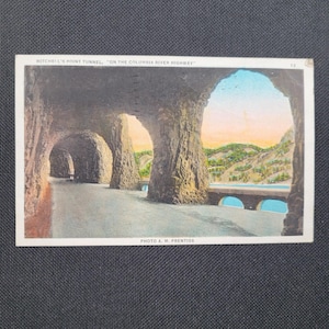 May include: Vintage postcard featuring Mitchell's Point Tunnel on the Columbia River Highway. The image shows a series of arched tunnels carved into the rock, with a road leading through them. The background reveals a bridge, river, and distant hills.