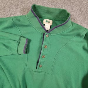 May include: Green long-sleeve shirt with a button-up neckline and a purple trim on the collar and cuffs. The shirt has a relaxed fit and is made of a soft material. The REI logo is visible on the tag. This shirt is perfect for casual wear.