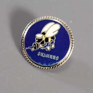 May include: A circular metal pin featuring a dark blue enamel background with a silver-toned rope-style border. The center displays a white and black bee graphic holding a small object, with the text SIEMENS printed in silver lettering below the bee.