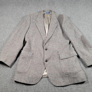 May include: A gray tweed blazer with a notched lapel, two front pockets, and a suede elbow patch. The blazer has a two-button closure and a light-colored lining. The fabric has flecks of red and brown.