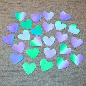 May include: A collection of heart-shaped paper cutouts in shades of purple, green, and white. The hearts are arranged on a textured, brown surface. The colors blend together, creating a gradient effect. The hearts are a craft or decorative item.
