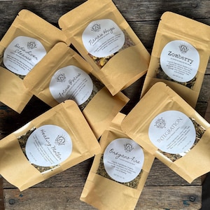 Puede incluir: Seis bolsas de papel marrón que contienen mezclas de infusiones, cada una con una etiqueta circular blanca. Las etiquetas muestran el logotipo de la marca y los nombres: Dad's Liver Cleanse, Nana's Hugs, Zomberry, Focus Pocus, Healing Mother, Oregano-Fire e INTEGRATION.