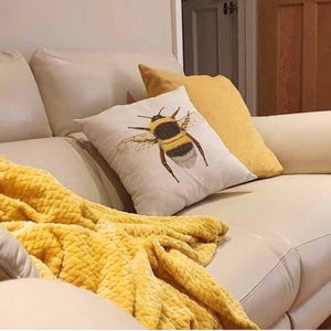K&ouml;nnte beinhalten: Cremefarbene Couch mit gelber Decke und Kissen. Ein Kissen zeigt eine detaillierte Illustration einer Hummel. Ein weiteres Kissen ist einfarbig gelb. Die Decke ist strukturiert, goldgelb. Gem&uuml;tliche Wohnatmosph&auml;re.