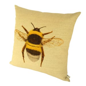 K&ouml;nnte beinhalten: Ein quadratisches, hellgelbes Kissen mit einer detaillierten Illustration einer Hummel. Die Hummel hat einen schwarz-gelb gestreiften K&ouml;rper und durchscheinende Fl&uuml;gel. Das Kissen hat ein strukturiertes Aussehen. Die Hummel ist mit ausgebreiteten Fl&uuml;geln dargestellt.