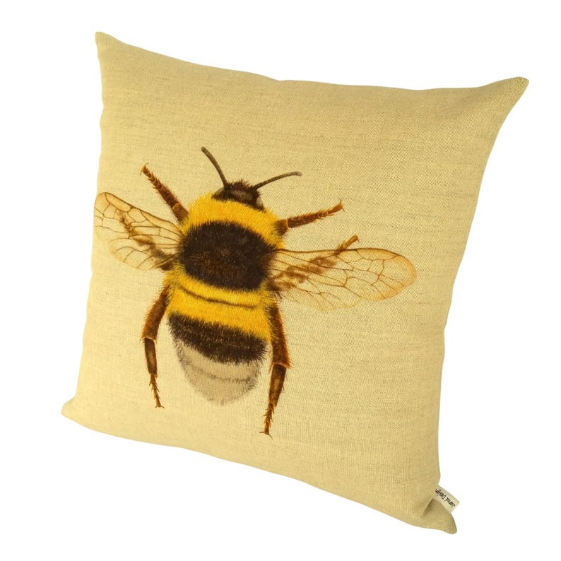 K&ouml;nnte beinhalten: Ein quadratisches, hellgelbes Kissen mit einer detaillierten Illustration einer Hummel. Die Hummel hat einen schwarz-gelb gestreiften K&ouml;rper und durchscheinende Fl&uuml;gel. Das Kissen hat ein strukturiertes Aussehen. Die Hummel ist mit ausgebreiteten Fl&uuml;geln dargestellt.