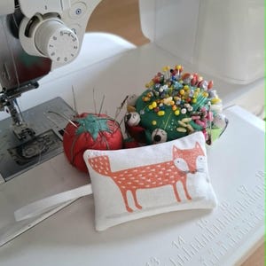 May include: A rectangular white fabric pincushion featuring an orange fox illustration, positioned next to a red tomato-shaped pincushion and a green round pincushion filled with multicoloured sewing pins, all resting on a white sewing machine.