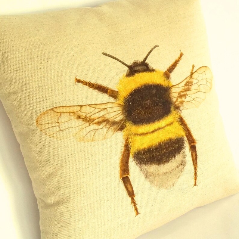K&ouml;nnte beinhalten: Ein quadratisches, cremefarbenes Zierkissen mit einer detaillierten Illustration einer Hummel. Die Hummel hat gelbe und schwarze Streifen, braune Beine und durchscheinende Fl&uuml;gel. Das Kissen hat ein strukturiertes Aussehen, das auf Leinen oder Baumwolle hindeutet.