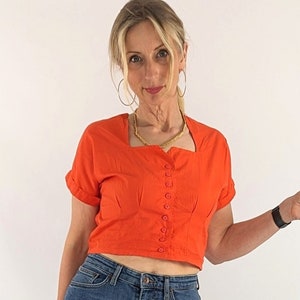 May include: A bright orange top with a square neckline and button detailing, worn with blue denim jeans. The outfit is accessorised with a gold necklace and large hoop earrings.
