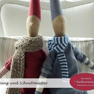 Könnte beinhalten: Zwei handgemachte Weihnachtswichtelpuppen, eine trägt ein rotes Kleid und einen braunen Strick Schal, die andere trägt ein blaues Kleid und einen weiß-blau gestreiften Schal. Die Puppen sitzen auf einem weiß-braun gestreiften Kissen. Der Text "Weihnachts-Wichtel" ist im Hintergrund sichtbar.