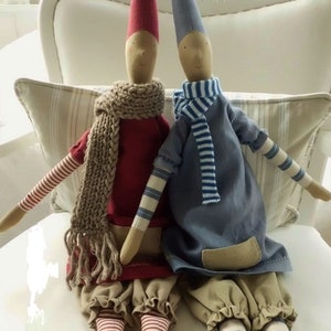 May include: Two handmade fabric dolls with pointed hats, one wearing a red dress with a brown scarf and the other wearing a blue dress with a white and blue striped scarf. Both dolls have striped legs and are sitting on a white chair.