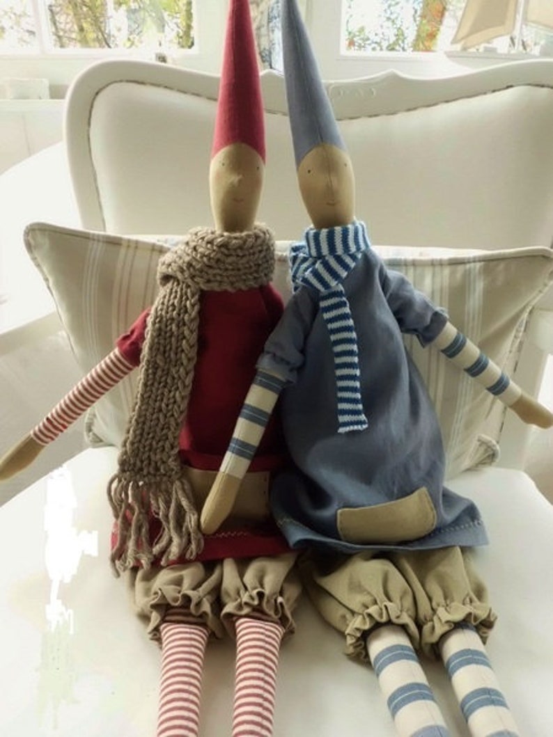 May include: Two handmade fabric dolls with pointed hats, one wearing a red dress with a brown scarf and the other wearing a blue dress with a white and blue striped scarf. Both dolls have striped legs and are sitting on a white chair.