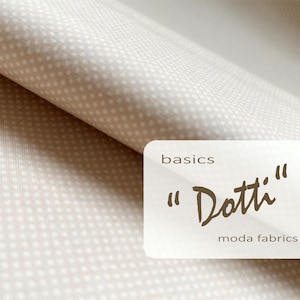 May include: Beige fabric with a small white polka dot pattern. The fabric is folded, showing texture and depth. A white label with the text "basics" and "Dotti" in a handwritten style, along with "moda fabrics".