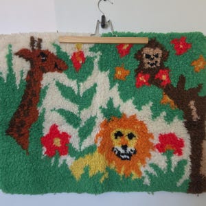 May include: A rectangular tufted wall hanging featuring a jungle scene with a brown giraffe, an orange lion, and a brown monkey in a tree. The design includes green foliage and red flowers on a white background, suspended from a wooden hanger.