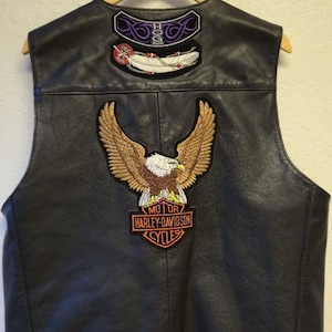 May include: A black leather vest featuring a large embroidered eagle patch above a Harley-Davidson Motor Cycles logo. The upper back displays two smaller patches, one with purple tribal designs and the text HOG 2004, and another with a white feather.