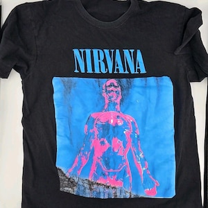 Puede incluir: Una camiseta negra de manga corta con la palabra NIRVANA en texto azul claro sobre un gráfico de una figura humana. La figura está representada en tonos rosa brillante y azul sobre un fondo azul, con una zona oscura y texturizada en la parte inferior.