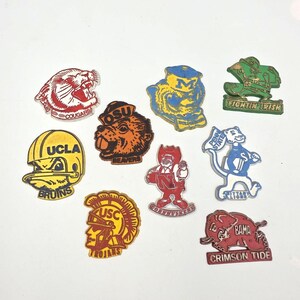 May include: A collection of nine colorful enamel pins featuring various university sports team mascots and logos, including UCLA Bruins, USC Trojans, OSU Beavers, Nebraska Cornhuskers, Penn State Nittany Lions, Alabama Crimson Tide, and Notre Dame Fighting Irish.