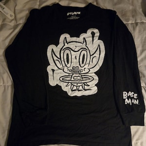 May include: A black long-sleeve t-shirt featuring a large white graphic of a cartoon devil character with horns and fangs on the chest. The text BASEMAN is printed in white on the left sleeve and inside the collar of the shirt.